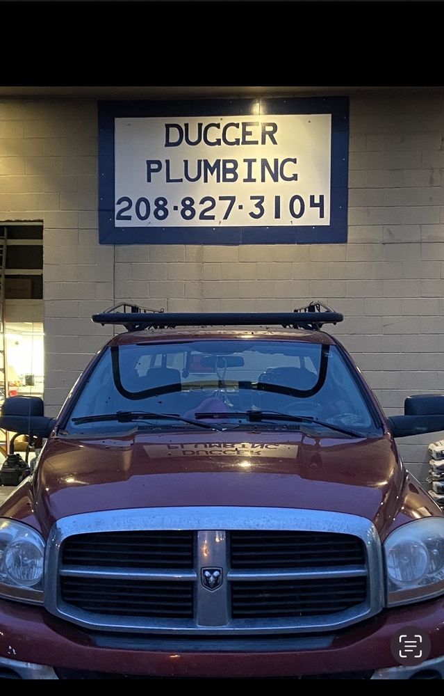 A maroon Dodge work van with roof racks is parked beneath a wall sign reading “Dugger Plumbing” and “208-827-3104,” giving a practical, local-business feel.
