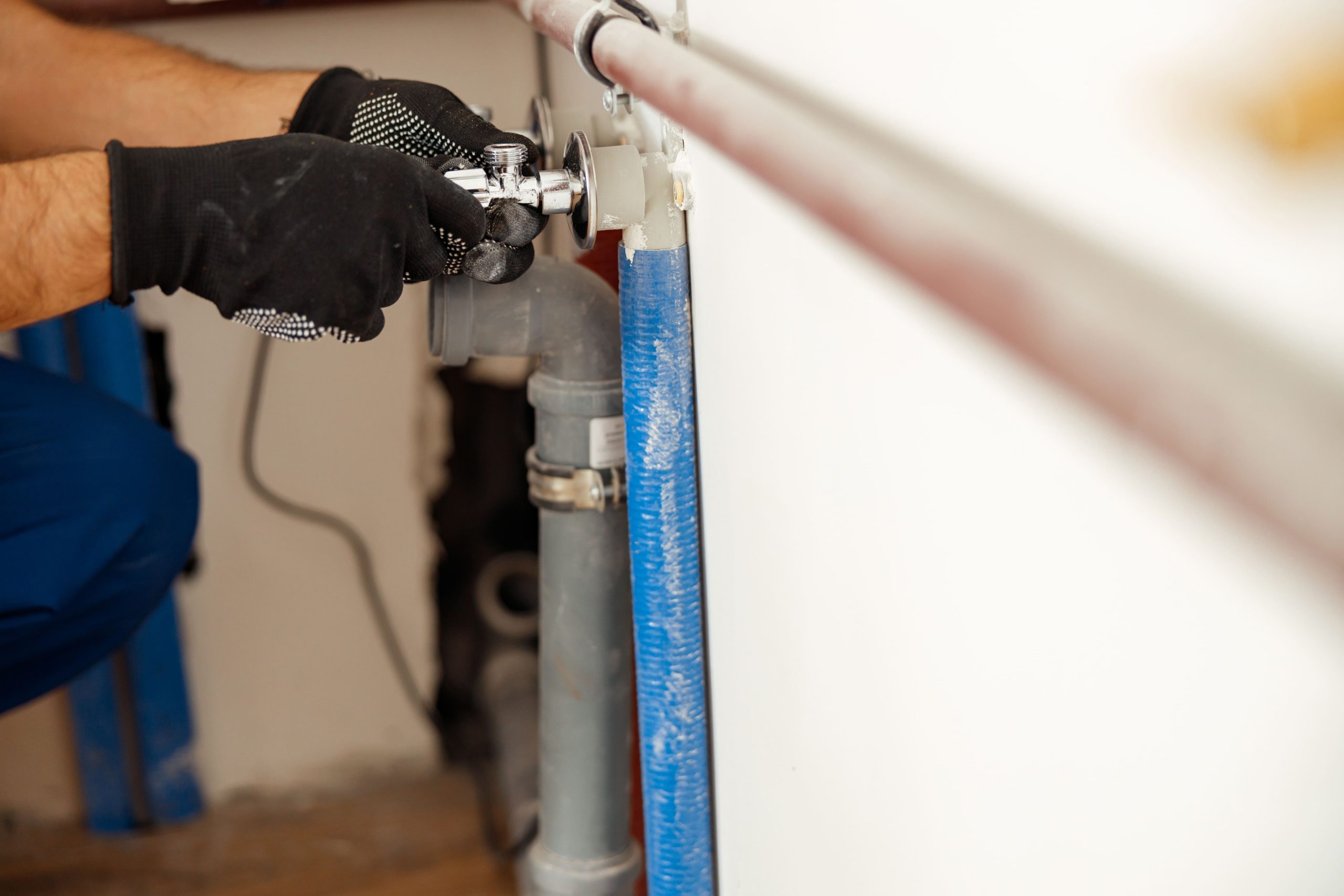 A plumber in black gloves adjusts a metal valve on a gray pipe beside a bright blue hose, working carefully in a home utility area.