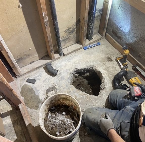 A worker kneels in a gutted bathroom beside a bucket of rubble and a rough floor opening, with exposed wall studs and power tools nearby.