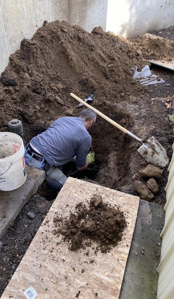A worker kneels in a narrow outdoor trench beside a large mound of soil, with a shovel, bucket, and dirt-covered board nearby; the scene feels laborious and practical.
