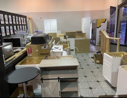 Crowded furniture showroom or storage room filled with stacked boxes, cabinets, tables, and appliances, creating a cluttered, busy atmosphere.